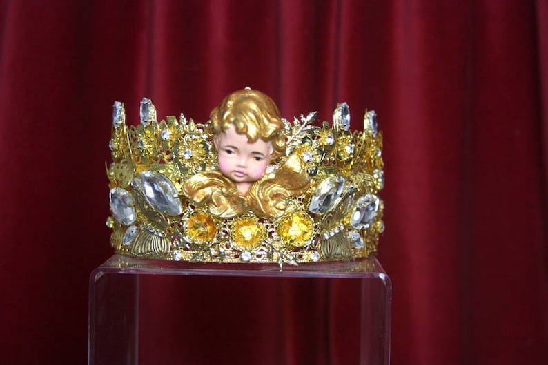 May include: A gold crown with clear rhinestones and a small porcelain cherub figurine in the center.