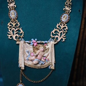 Peut inclure: Collier dor&eacute; orn&eacute; d'un ch&eacute;rubin central. Le collier comprend des accents de cam&eacute;e et un design d&eacute;taill&eacute; en volutes. Le ch&eacute;rubin est repr&eacute;sent&eacute; sur une banni&egrave;re avec des accents floraux. Une cha&icirc;ne d&eacute;licate avec un pendentif cam&eacute;e est suspendue en dessous.