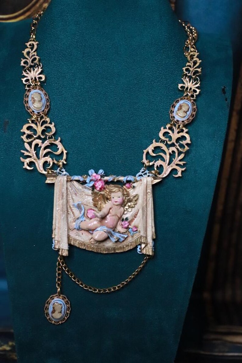 Peut inclure: Collier dor&eacute; orn&eacute; d'un ch&eacute;rubin central. Le collier comprend des accents de cam&eacute;e et un design d&eacute;taill&eacute; en volutes. Le ch&eacute;rubin est repr&eacute;sent&eacute; sur une banni&egrave;re avec des accents floraux. Une cha&icirc;ne d&eacute;licate avec un pendentif cam&eacute;e est suspendue en dessous.