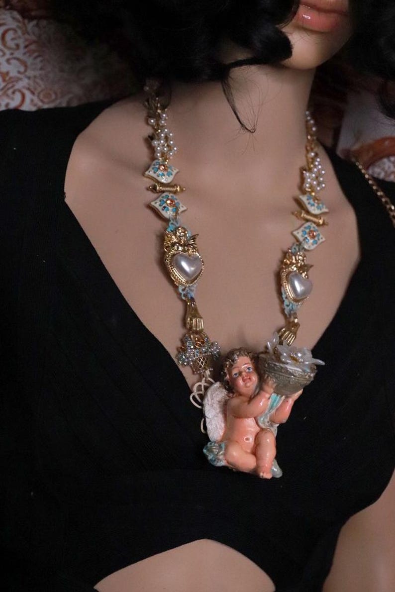 May include: A gold-toned necklace featuring a cherub holding a basket. The necklace has pearl and heart-shaped accents, with blue and white decorative elements. The cherub is pink and white, and the necklace is worn against a black top.
