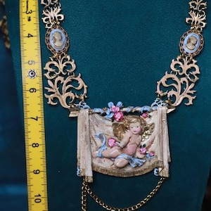 Peut inclure: Collier dor&eacute; orn&eacute; d'un pendentif ch&eacute;rubin et de cam&eacute;es. Le collier pr&eacute;sente un design d&eacute;taill&eacute; avec des volutes et une figure centrale de ch&eacute;rubin. Le collier mesure environ 25 cm de long.