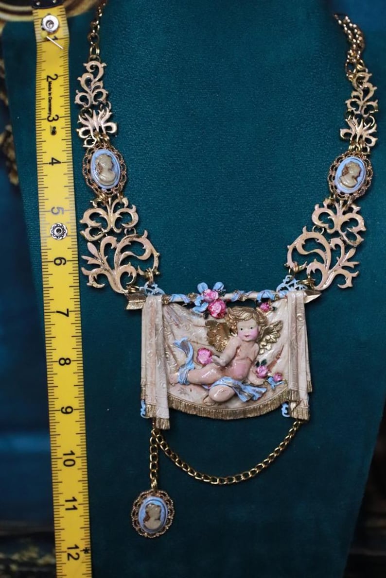 Peut inclure: Collier dor&eacute; orn&eacute; d'un pendentif ch&eacute;rubin et de cam&eacute;es. Le collier pr&eacute;sente un design d&eacute;taill&eacute; avec des volutes et une figure centrale de ch&eacute;rubin. Le collier mesure environ 25 cm de long.