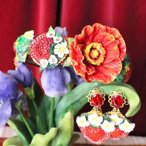 Puede incluir: Pendientes de amapola roja y fresa con flores blancas y gemas rojas. Los pendientes cuelgan de una planta verde con flores moradas.