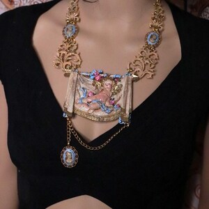 Peut inclure: Collier dor&eacute; orn&eacute; d'un pendentif rectangulaire central repr&eacute;sentant un ch&eacute;rubin, avec des d&eacute;tails bleus et roses. Le collier comprend une cha&icirc;ne avec un petit pendentif cam&eacute;e et des &eacute;l&eacute;ments de filigrane d&eacute;coratifs.