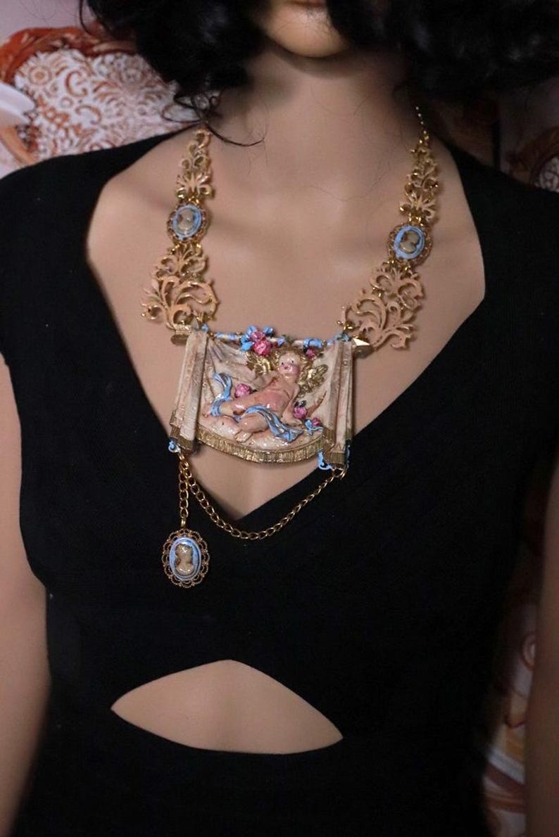 Peut inclure: Collier dor&eacute; orn&eacute; d'un pendentif rectangulaire central repr&eacute;sentant un ch&eacute;rubin, avec des d&eacute;tails bleus et roses. Le collier comprend une cha&icirc;ne avec un petit pendentif cam&eacute;e et des &eacute;l&eacute;ments de filigrane d&eacute;coratifs.