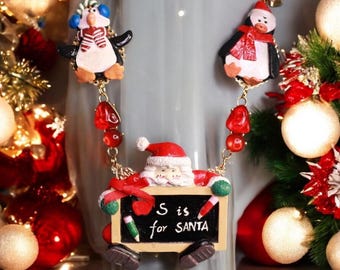 May include: A gold chain necklace with red beads and Christmas-themed charms. The charms include two penguins and a Santa Claus holding a chalkboard that says "S is for SANTA".