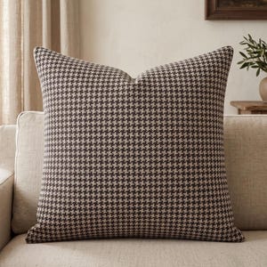 May include: A square throw pillow featuring a dark brown and beige houndstooth pattern. The fabric has a visible woven texture, and the pillow is plumped to maintain a structured, full shape with crisp, defined edges and corners.