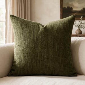 May include: A square throw pillow featuring a deep olive green fabric with a subtle, textured, woven pattern. The pillow has a structured, plump appearance with clean, straight edges and a soft, matte finish across the entire surface.