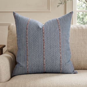 May include: A square throw pillow featuring a textured blue fabric with a subtle herringbone weave. The pillow is accented by three vertical stripes in a rust-orange and white color palette, running parallel from the top to the bottom edge.