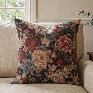 Könnte beinhalten: Ein quadratisches Zierkissen mit einem tiefen, dunkelburgunderfarbenen Hintergrund, verziert mit einem dichten, gewebten Gobelinmuster aus großen Rosen in Creme-, gedeckten Rottönen und Staubblau, durchsetzt mit grünen Blättern und kleinen Blüten.