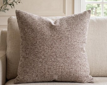 Op de afbeelding: Een vierkant sierkussen met een getextureerde, geweven stof in een gevarieerde mix van lichtbruine, beige en donkerbruine draden. Het kussen heeft een consistent, dicht weefpatroon over het hele oppervlak, wat zorgt voor een neutrale, aardse uitstraling.