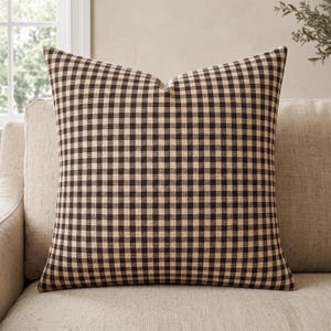 May include: A square throw pillow featuring a classic dark brown and beige gingham check pattern. The fabric displays a consistent, repeating grid design across the entire surface of the pillow, creating a balanced and uniform appearance throughout.