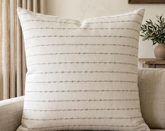 May include: A square throw pillow featuring an off-white fabric base with horizontal rows of textured, light brown stitched lines. The pillow has a woven appearance with a consistent pattern of thin, raised stripes running across the entire surface.