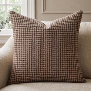 May include: A square throw pillow featuring a classic brown and beige houndstooth pattern. The fabric has a woven texture, and the pillow is filled to maintain a plump, structured shape. The design is consistent across the entire front surface.
