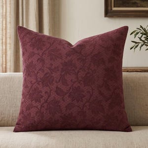 May include: A square throw pillow in a deep burgundy color featuring a raised, tonal floral pattern with intricate leaf and petal details. The fabric has a subtle, textured lattice background that complements the overall dark red floral design.