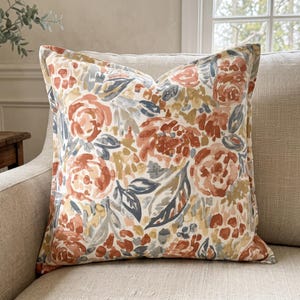 May include: A square throw pillow featuring a watercolor-style floral pattern in shades of terracotta, muted blue, and mustard yellow on a cream background. The design consists of abstract flower blossoms and leafy foliage across the fabric surface.