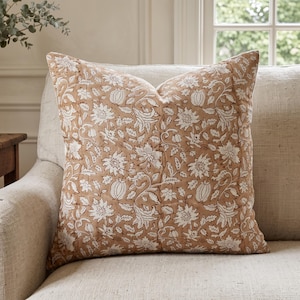 May include: A square throw cushion featuring a tan background with a repeating white floral and botanical pattern. The fabric has visible quilted stitching lines running vertically across the surface, creating a textured appearance on the cushion.