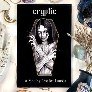 May include: A black and gold zine titled "cryptic" with a drawing of a woman in a coffin. The text "a zine by Jessica Lauser" is printed below the image.