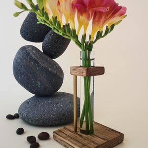 May include: A wooden stand with a glass test tube vase holding a bouquet of pink and yellow freesia flowers. The stand is sitting on a white surface with gray stones and coffee beans.