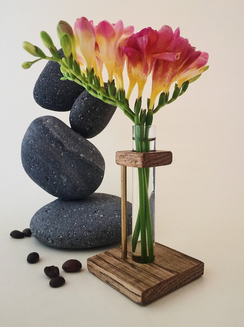 May include: A wooden stand with a glass test tube vase holding a bouquet of pink and yellow freesia flowers. The stand is sitting on a white surface with gray stones and coffee beans.