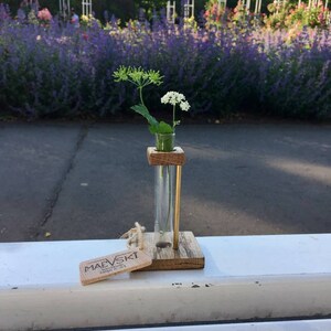 May include: A small wooden vase with a glass test tube insert holds a sprig of white flowers. The vase is sitting on a white surface with a wooden tag that reads "MAEVSKI Handmade with Love" in front of it.