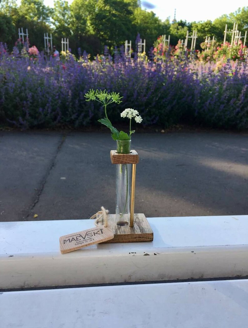May include: A small wooden vase with a glass test tube insert holds a sprig of white flowers. The vase is sitting on a white surface with a wooden tag that reads "MAEVSKI Handmade with Love" in front of it.