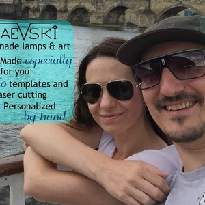 May include: A couple smiles for a photo in front of a bridge. The man is wearing a green baseball cap and sunglasses. The woman is wearing sunglasses. The text on the image reads "MAEVSKİ handmade lamps & art Made especially for you No templates and laser cutting Personalized by hand".
