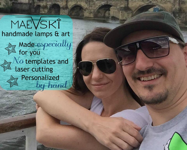 May include: A couple smiles for a photo in front of a bridge. The man is wearing a green baseball cap and sunglasses. The woman is wearing sunglasses. The text on the image reads "MAEVSKİ handmade lamps & art Made especially for you No templates and laser cutting Personalized by hand".