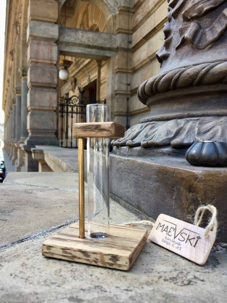 May include: A wooden test tube holder with a gold metal rod and a clear glass test tube. The holder is sitting on a stone surface with a tag that reads "Maevski Handmade Lamps & Art".