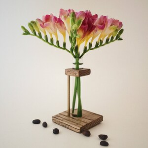 May include: A wooden flower vase with a glass test tube insert. The vase is made of rustic wood and has a square base. The test tube is filled with pink and yellow freesia flowers.