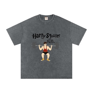 Pode incluir: T-shirt cinzento escuro com o texto "Harry Squatter and the Squats Rock of Mysteries". O gráfico mostra uma figura de desenho animado em agachamento com uma barra. A camisa tem gola redonda e mangas curtas.
