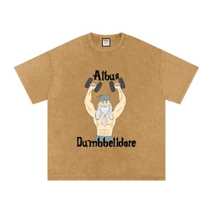 Puede incluir: Camiseta de color mostaza con una ilustración de dibujos animados de un mago musculoso levantando pesas. El texto encima del mago dice "Albus", y debajo, "Dumbbelldore". La camiseta tiene cuello redondo y mangas cortas.