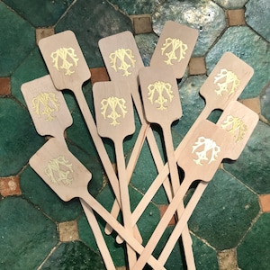 May include: A collection of wooden drink stirrers with a light wood finish. Each stirrer features a rectangular paddle with a gold-colored monogram design. The stirrers are arranged on a green and brown tiled surface.