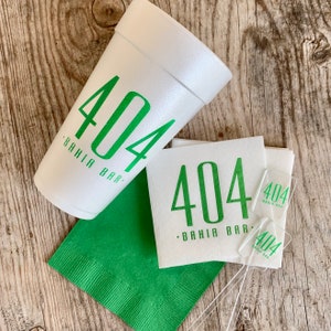 May include: A white foam cup, a stack of white paper napkins, a single green paper napkin, and two clear plastic stirrers. The cup, napkins, and stirrers all feature the green printed text 404 BAHIA BAR in a clean, sans-serif font style.