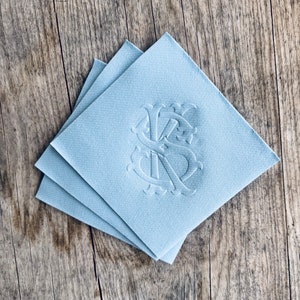 May include: Three light blue square napkins with an embossed monogram design. The monogram features intertwined letters, possibly initials, in a decorative script. The napkins are stacked on a wooden surface, creating a textured contrast.