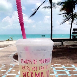 May include: A clear plastic cup with a pink straw, filled with a yellow beverage. The cup has a white lid and text that reads "Always be yourself unless you can be a mermaid or Chelsea Stelte" in pink. A mermaid graphic is also visible.