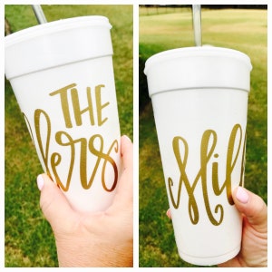 May include: A white foam cup with a metallic gold script design. The cup features the text THE and a cursive script below it, with a silver straw inserted through the lid. The design is displayed across two views of the same cup.