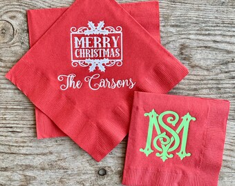 May include: Red paper napkins with white text that reads "MERRY CHRISTMAS" and "The Carsons". A second napkin has a green monogram. The napkins are on a wooden surface.
