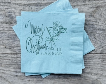 May include: A stack of light blue cocktail napkins with a printed design. The design includes the words "Merry Christmas" in cursive, a martini glass with floral accents, and the name "The Carsons".