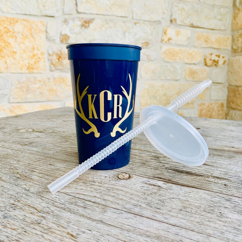 May include: A navy blue tumbler with a gold antler design and the initials "KCR". The tumbler includes a clear plastic lid and a matching straw. The cup is sitting on a wooden surface.