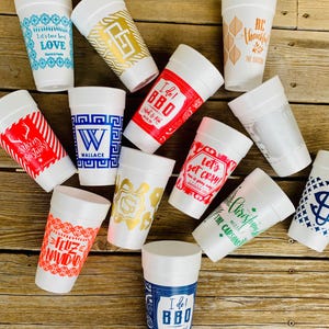 May include: A collection of white foam cups with various designs and text. Some cups feature holiday greetings like "Merry Christmas" and "Feliz Navidad." Others have phrases like "Let's get CRAY!" and "I do! BBQ." The cups are decorated with blue, red, and gold accents.
