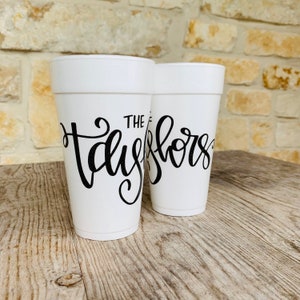 May include: Two white foam cups featuring black cursive lettering. The front cup displays the text "THE TAYLORS" in a stylized script font, while the second cup behind it shows a partial view of the same black cursive text design.