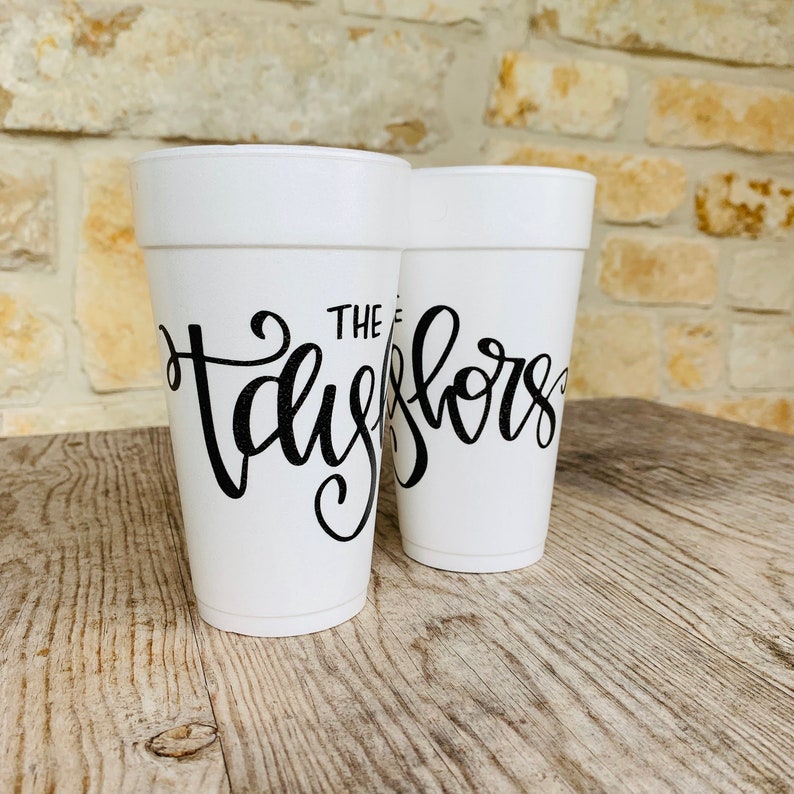 May include: Two white foam cups featuring black cursive lettering. The front cup displays the text "THE TAYLORS" in a stylized script font, while the second cup behind it shows a partial view of the same black cursive text design.