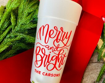 May include: A white foam cup featuring red text that reads Merry and Bright and THE CARSONS in a script and sans-serif font. The cup is positioned against a backdrop of green pine needles and a wide, bright red velvet ribbon.