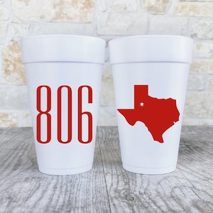 May include: Two white foam cups. One cup has the number "806" in red. The other cup has a red Texas state outline with a white star. The cups are on a wooden surface.