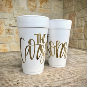 May include: Two white foam cups with gold lettering that reads "The Carsons". The cups are sitting on a wooden surface, with a stone wall in the background. The cups are a light, off-white color.