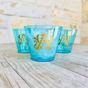 May include: Three turquoise plastic cups with gold lettering. The cups have the text "ALLISON & WILLIAM" below a gold monogram. The cups are on a wooden surface with a light-colored stone background.