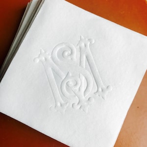 May include: A stack of white paper napkins with an embossed monogram design. The monogram features the letters "S" and "M" in an elegant, swirling font. The napkins are stacked on a reddish-brown surface.