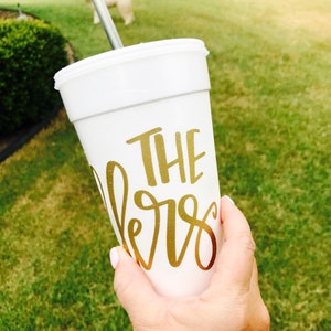 May include: A white styrofoam cup featuring gold metallic lettering that reads THE lers in a combination of block and script fonts. The cup includes a white lid and a thin silver metal straw extending from the top, designed for holding cold beverages.