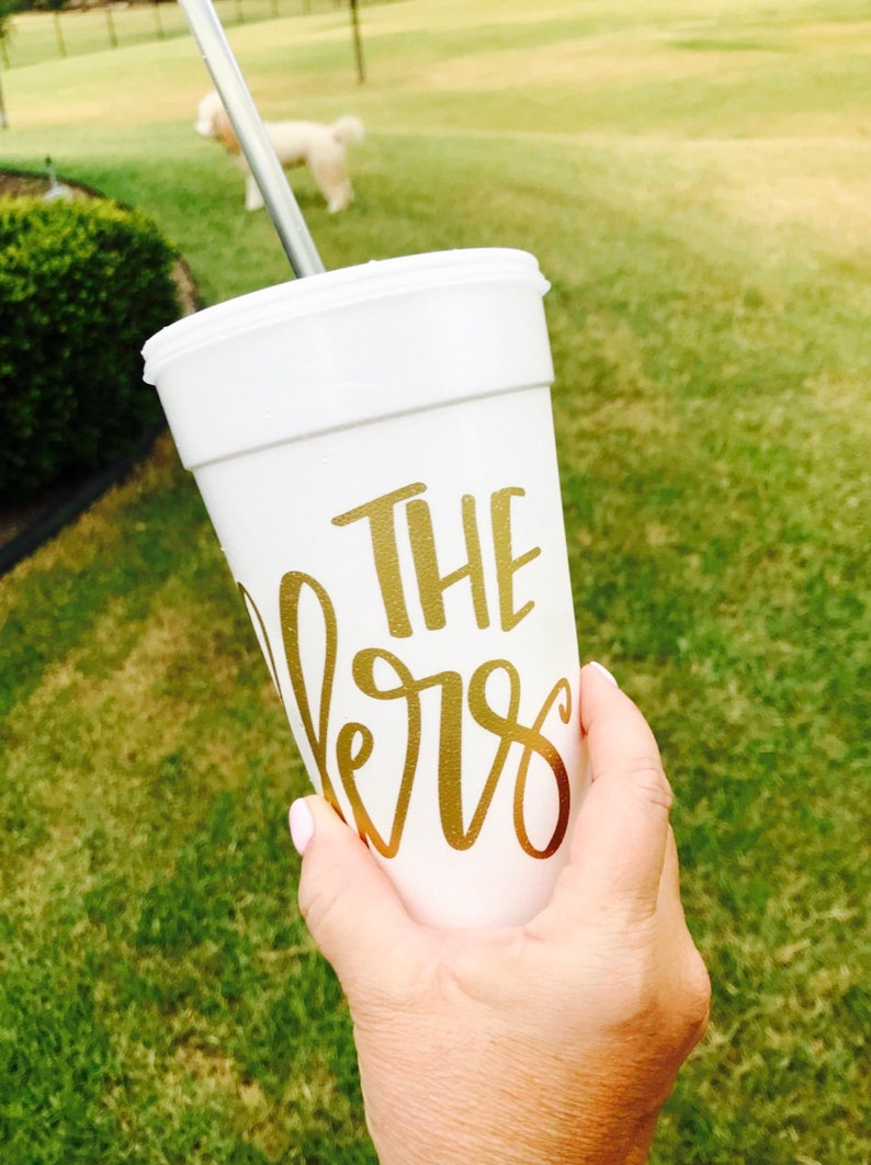 May include: A white styrofoam cup featuring gold metallic lettering that reads THE lers in a combination of block and script fonts. The cup includes a white lid and a thin silver metal straw extending from the top, designed for holding cold beverages.